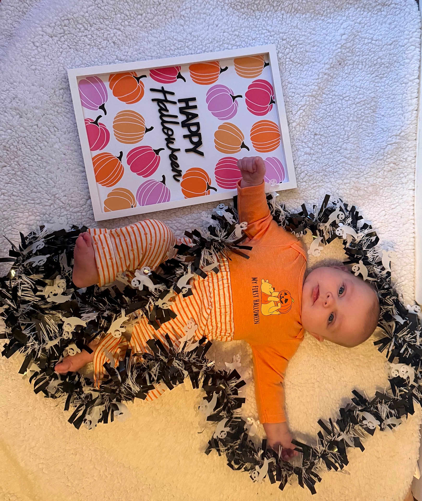 Aiyanna is registered to the contest to win money with this photo: baby, infant, halloween, orange_clothing, striped_pants, blanket, garland, ghosts, pumpkins, sign, holiday, festive, cute, lying_down, indoors, decorations, child, celebration, smiling, portrait