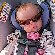 Scarlett is registered to the contest to win money with this photo: baby, infant, child, car_seat, safety_harness, sunglasses, pacifier, car, vehicle, seated, portrait, face, head, clothing, pink, black, indoor, travel, passenger, safety_warning