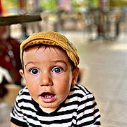 Manil a rejoint le concours — aidez-le/la à gagner de superbes lots ! toddler, child, striped_shirt, yellow_cap, surprised_expression, blue_eyes, hands, table, outdoor, blurred_background, greenery, portrait, casual_clothing, young_child, curious, face, person, daylight, candid, closeup