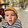 toddler, child, striped_shirt, yellow_cap, surprised_expression, blue_eyes, hands, table, outdoor, blurred_background, greenery, portrait, casual_clothing, young_child, curious, face, person, daylight, candid, closeup