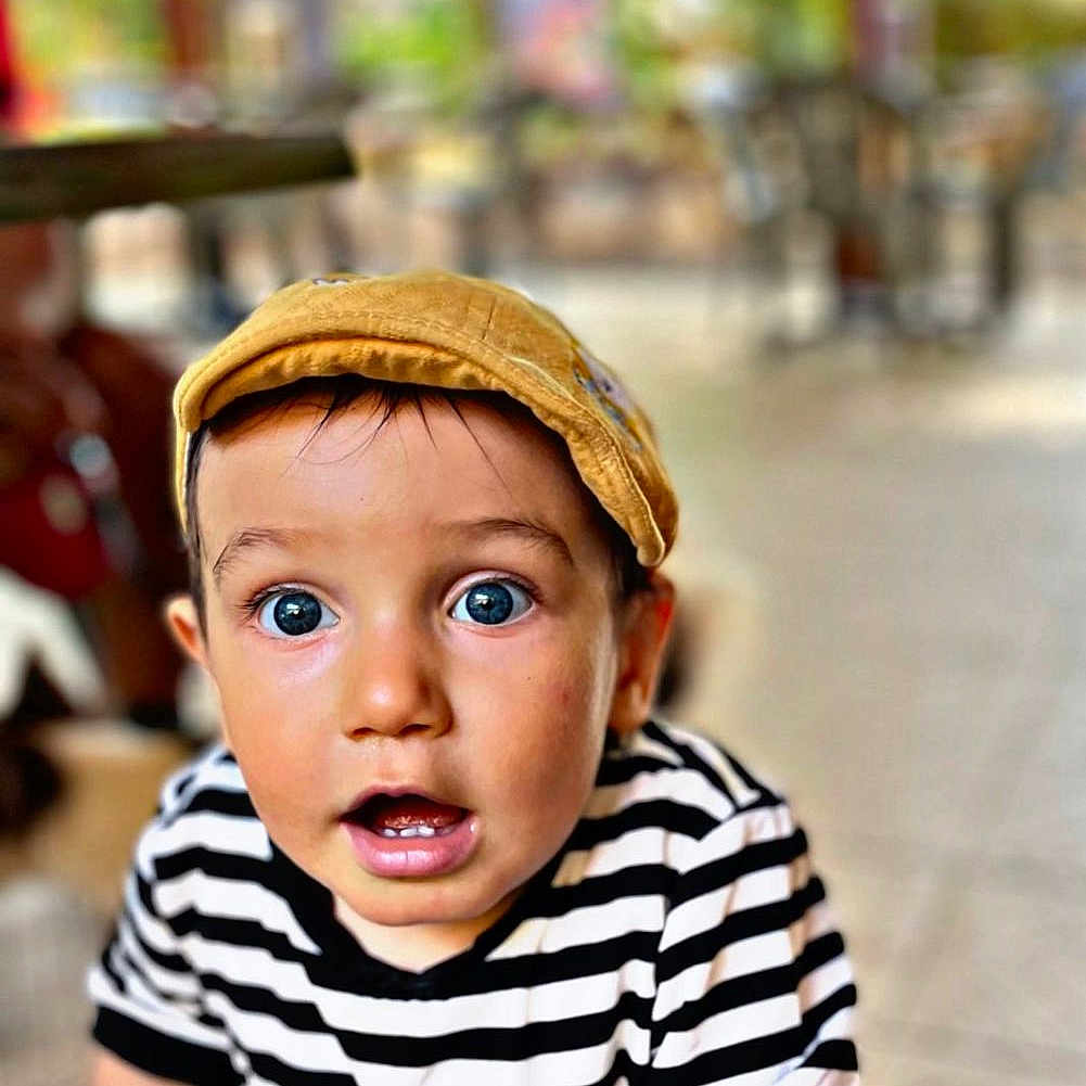 Manil a rejoint le concours — aidez-le/la à gagner de superbes lots ! blue_eyes, blurred_background, candid, casual_clothing, child, closeup, curious, daylight, face, greenery, hands, outdoor, person, portrait, striped_shirt, surprised_expression, table, toddler, yellow_cap, young_child