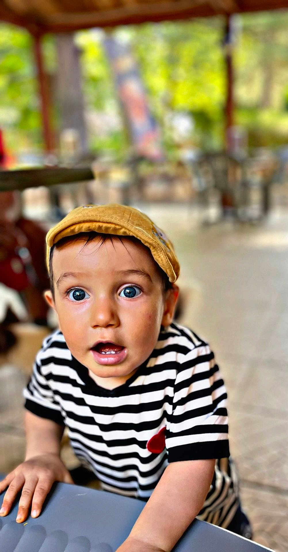 Manil a rejoint le concours — aidez-le/la à gagner de superbes lots ! toddler, child, striped_shirt, yellow_cap, surprised_expression, blue_eyes, hands, table, outdoor, blurred_background, greenery, portrait, casual_clothing, young_child, curious, face, person, daylight, candid, closeup