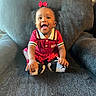 baby, child, smiling, red_dress, bow, shoes, armchair, happy, tongue_out, cute, indoor, portrait, person, sitting, infant, black_hair, clothing, playful, young_child, cozy