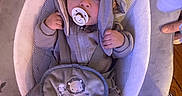 Micah is registered to the contest to win money with this photo: baby, pacifier, hood, baby_seat, gray_clothing, socks, plush, infant, cute, child, indoor, fabric, pouch, hand, floor, pattern, toy, soft, seated, curious