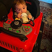Micah is registered to the contest to win money with this photo: baby, bottle, toy_car, jeep, indoor, carpet, floor, child, milk, seat, hand, face, eyes, clothing, furniture, play, vehicle, cute, infant, sitting