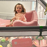 Rose joined the competition — help win amazing prizes! toddler, child, pink_clothing, plush_chair, playpen, toys, mesh, indoor, window, natural_light, smiling, happy, person, young_child, soft_texture, home, sitting, colorful, cozy, daylight