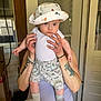 adult_hands, baby, casual, child, clothing, curious_expression, dinosaur_hat, dinosaur_shorts, door, hat, holding, indoor, light_skin, person, rings, shorts, socks, standing, tattoos