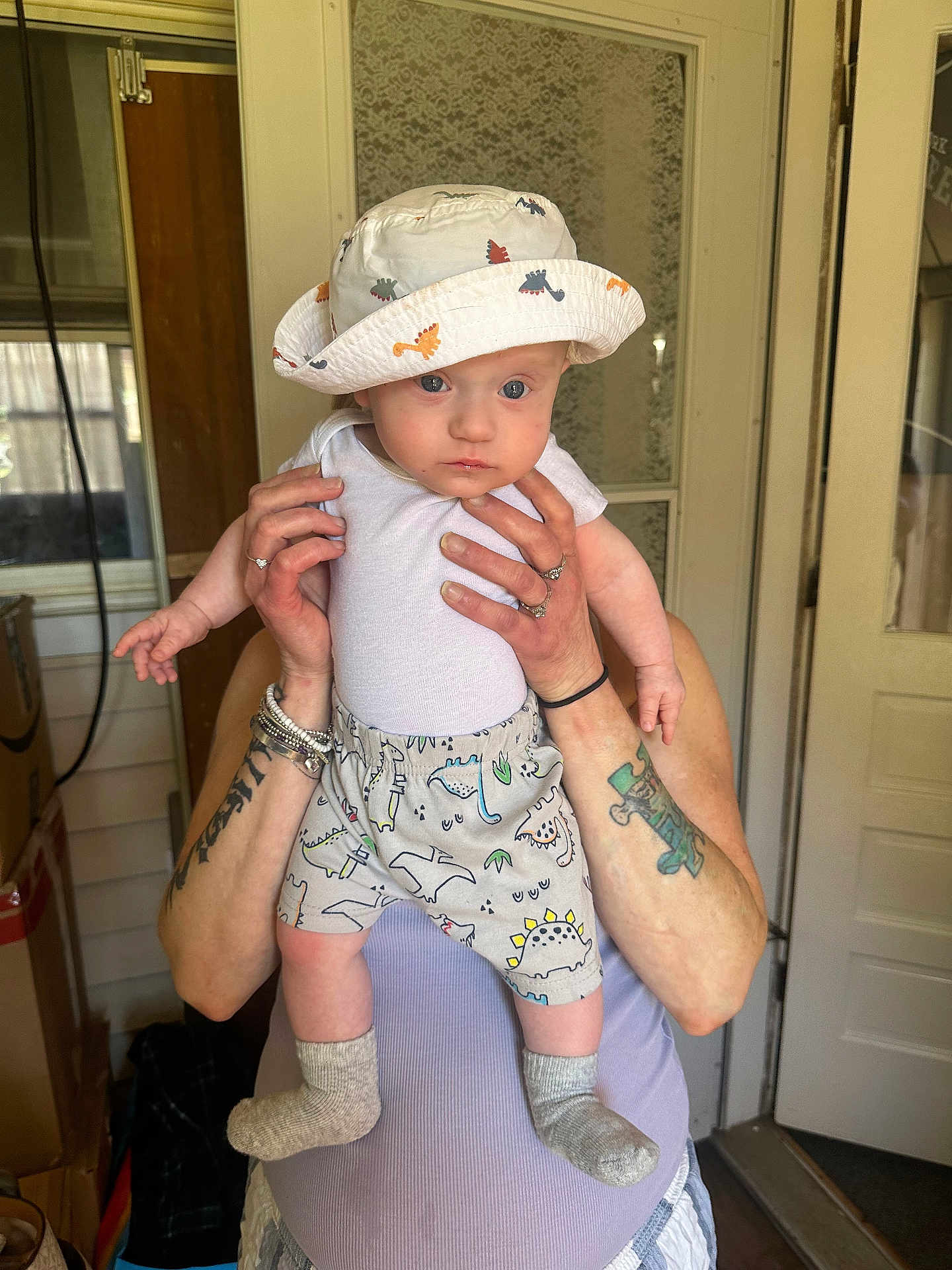 Maverick joined the competition — help win amazing prizes! baby, child, dinosaur_hat, dinosaur_shorts, socks, tattoos, rings, adult_hands, indoor, door, curious_expression, holding, person, clothing, shorts, hat, standing, light_skin, casual