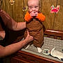adult_arm, animal_stickers, baby, blanket, changing_table, child, cute, expression, furniture, hand_support, home, indoor, orange_clothing, overalls, person, portrait, socks, toddler, toy, wood_paneling