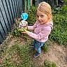 child, girl, outdoor, grass, greenery, fence, pink_hoodie, jeans, shoes, package, owl_package, playful, curious, nature, grass_patch, dirt, casual_clothing, young_child, person, standing
