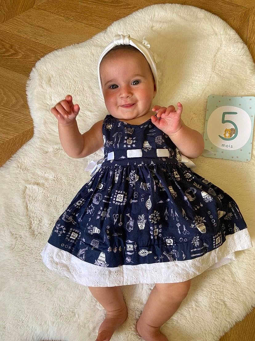 Lola a rejoint le concours — aidez-le/la à gagner de superbes lots ! baby, infant, smiling, dress, headband, white_rug, wooden_floor, milestone_card, 5_months, cute, child, happy, lying_down, indoors, person, portrait, hands_up, skin, face, eyes