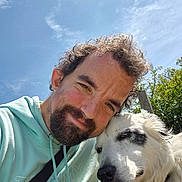 Marley is registered to the contest to win money with this photo: beard, black_patch, casual_clothing, close_up, clouds, curly_hair, dog, friendship, greenery, happy, hoodie, man, nature, outdoor, pet, portrait, sky, smiling, sunlight, white_dog