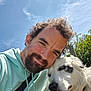 beard, black_patch, casual_clothing, close_up, clouds, curly_hair, dog, friendship, greenery, happy, hoodie, man, nature, outdoor, pet, portrait, sky, smiling, sunlight, white_dog