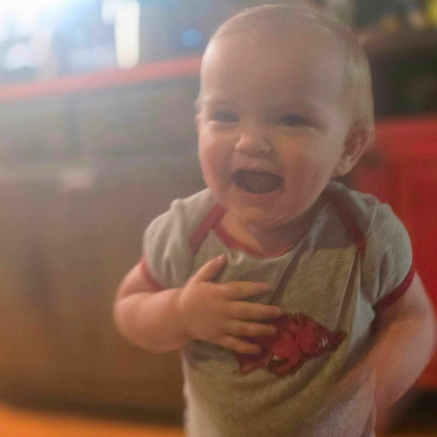 Stevie joined the competition — help win amazing prizes! baby, child, infant, smiling, laughing, indoor, portrait, face, person, standing, hand, shirt, clothing, red, gray, furniture, blurred_background, warm_lighting, candid, happy