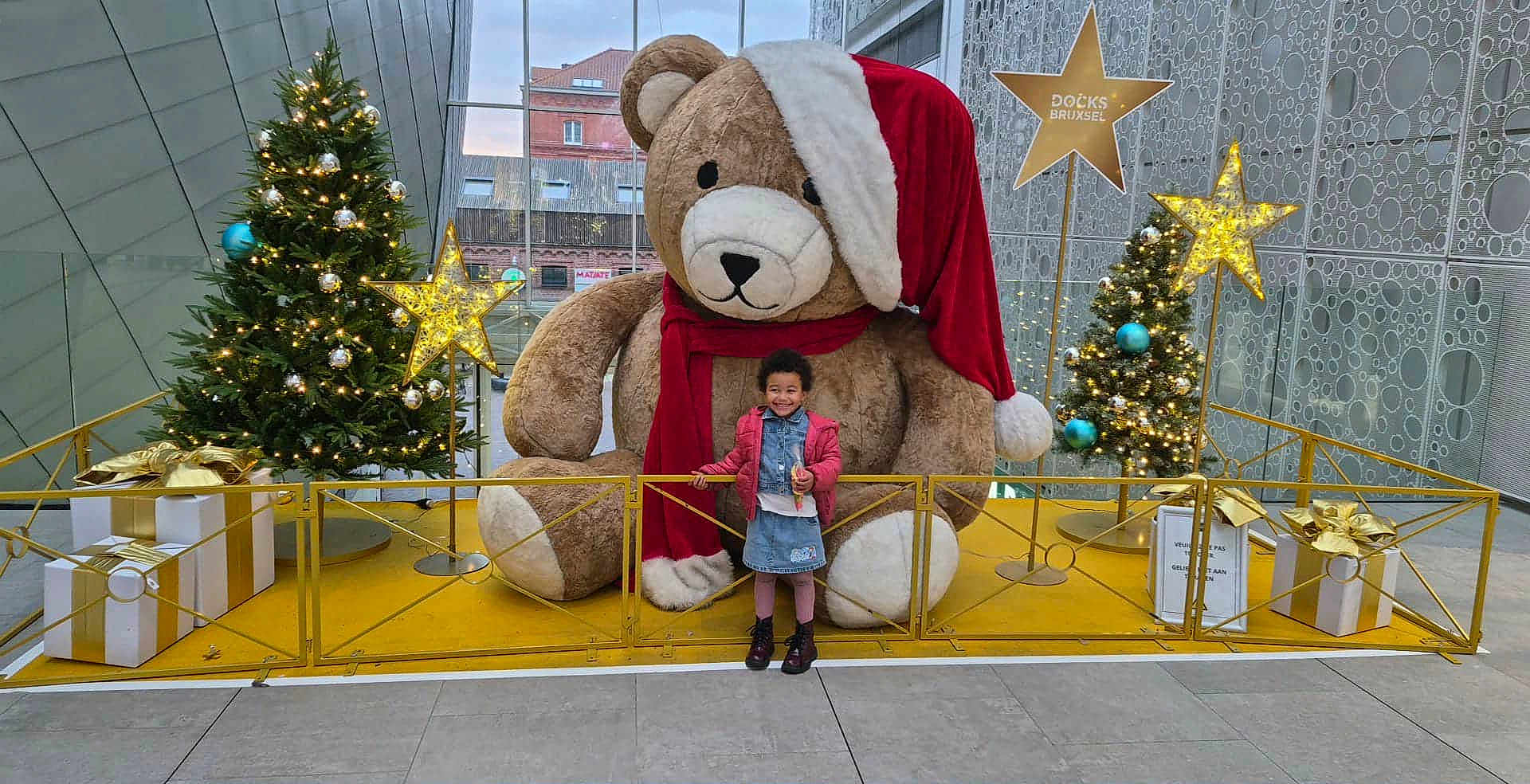 Maelys Maes Leonard participe au concours pour gagner de l'argent avec cette photo : child, teddy_bear, christmas_tree, holiday_decor, gift_box, indoor, festive, lights, smiling, winter_clothing, scarf, santa_hat, modern_architecture, glass_ceiling, floor_tiles, metal_railing, happy, posing, decorations, celebration