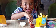 Maelys Maes Leonard a rejoint le concours — aidez-le/la à gagner de superbes lots ! child, smiling, curly_hair, food, fries, dipping_sauce, table, sitting, indoor, can, nugget, happy, person, restaurant, tray, plastic_container, clothing, beverage, fast_food, casual