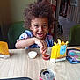 child, smiling, curly_hair, food, fries, dipping_sauce, table, sitting, indoor, can, nugget, happy, person, restaurant, tray, plastic_container, clothing, beverage, fast_food, casual