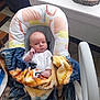 baby, blanket, baby_seat, indoor, table, cup, wood_coaster, floor_tiles, person, face, clothing, hand, blanket_pattern, seatbelt, expression, head, furniture, colorful, curious, sitting