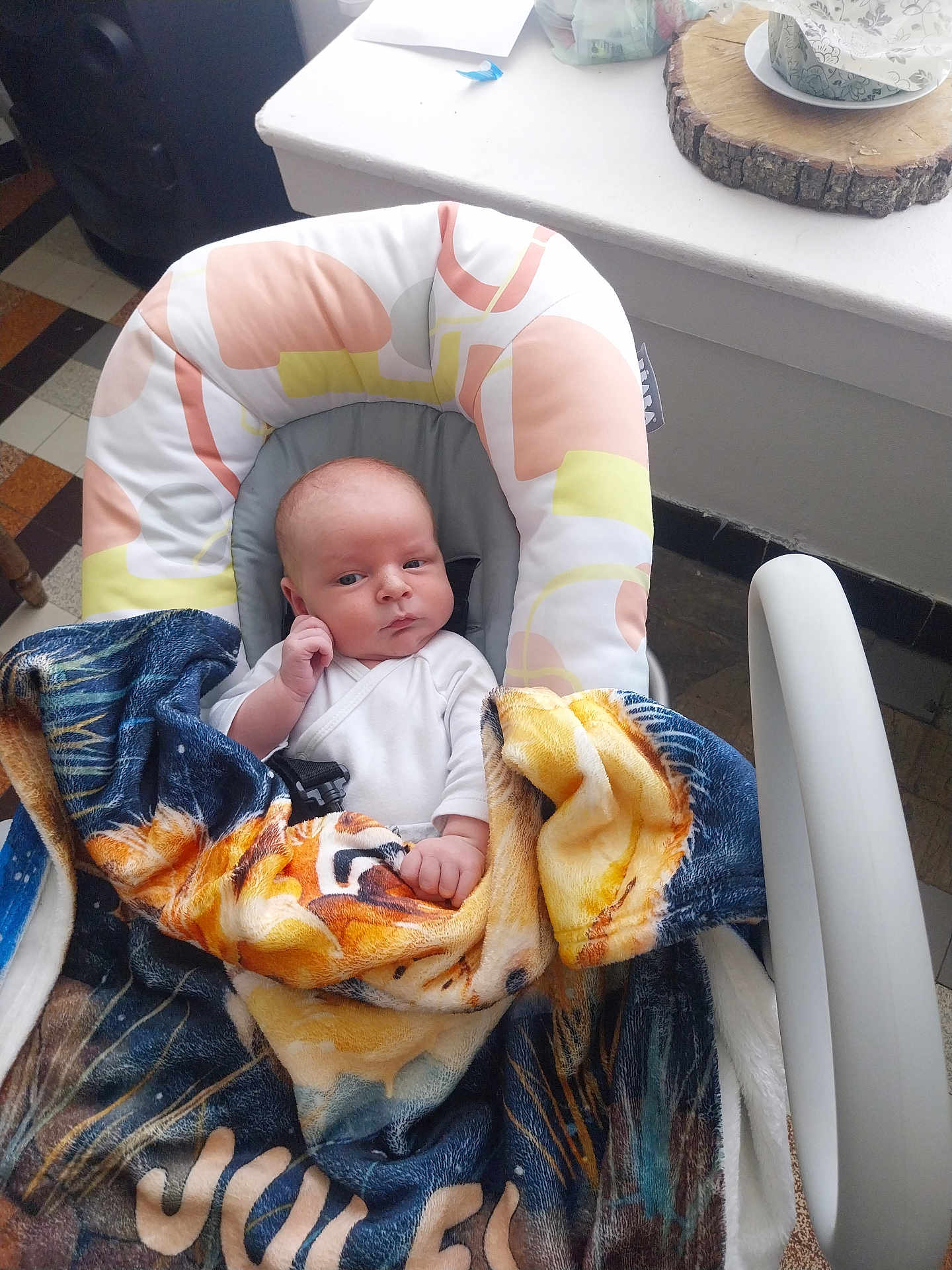 Jules a rejoint le concours — aidez-le/la à gagner de superbes lots ! baby, blanket, baby_seat, indoor, table, cup, wood_coaster, floor_tiles, person, face, clothing, hand, blanket_pattern, seatbelt, expression, head, furniture, colorful, curious, sitting
