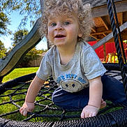 Logan is registered to the contest to win money with this photo: toddler, child, curly_hair, blue_eyes, swing, rope_swing, outdoor, playground, play_structure, slide, grass, sunny, smiling, grey_tshirt, navy_pants, happy, daytime, backyard, person, fun