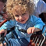 child, toddler, curly_hair, smiling, shopping_cart, blue_shirt, adult, glasses, outdoor, sunlight, happy, person, playful, metal, hand, face, jeans, daylight, closeup, family