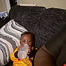 Legacy is registered to the contest to win money with this photo: baby, bottle, couch, child, person, indoor, clothing, yellow_outfit, relaxing, curious, dark_gray_couch, pillow, striped_pillow, hand, sitting, feeding, young_child, infant, casual, home
