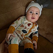 Bea joined the competition — help win amazing prizes! baby, child, onesie, hat, couch, drool, hand, footwear, black_boots, brown_furniture, curious_expression, indoor, person, infant, seated, cute, wearing, colorful_clothing, portrait, cozy