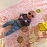 baby, infant, sunglasses, crib, pink, floral, toy, stuffed_animal, baby_clothing, pants, long_sleeve_shirt, lying_down, indoor, child, cute, colorful, soft_toy, accessory, headwear, person