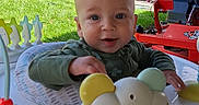 Atlas is registered to the contest to win money with this photo: baby, child, infant, smiling, outdoor, activity_center, toy, playtime, red_truck, truck, flowers, hanging_basket, grass, backyard, sunlight, portrait, sitting, green_clothing, baby_gear, colorful