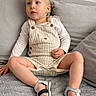 toddler, child, blonde_hair, overalls, sandals, long_sleeve_shirt, couch, gray_couch, indoor, sitting, curious, young_child, casual_clothing, footwear, comfortable, relaxed, home_interior, furniture, portrait, person