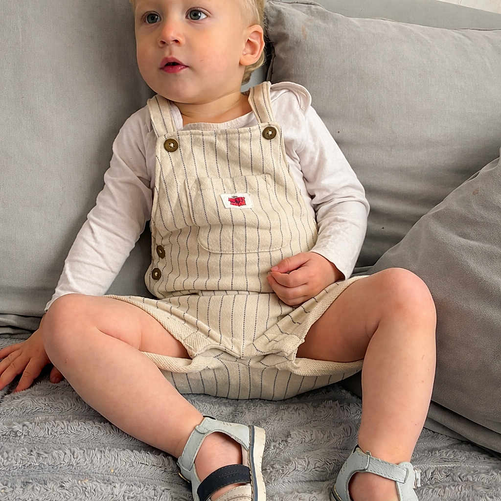 Neylan participe au concours pour gagner de l'argent avec cette photo : toddler, child, blonde_hair, overalls, sandals, long_sleeve_shirt, couch, gray_couch, indoor, sitting, curious, young_child, casual_clothing, footwear, comfortable, relaxed, home_interior, furniture, portrait, person