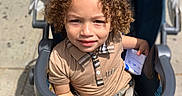 Andres joined the competition — help win amazing prizes! child, toddler, stroller, curly_hair, toy, outdoor, sunlight, sidewalk, beige_shirt, baby, happy, person, seat_belt, hand, paper, face, portrait, summer, casual_clothing, urban