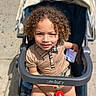 child, toddler, stroller, curly_hair, toy, outdoor, sunlight, sidewalk, beige_shirt, baby, happy, person, seat_belt, hand, paper, face, portrait, summer, casual_clothing, urban
