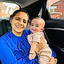 woman, baby, child, car_interior, blue_clothing, pink_clothing, smile, happy, portrait, holding, seat, window, urban_background, family, affection, person, casual, daylight, skin, hand