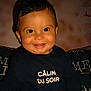 baby, background_blur, black_hair, child, closeup, couch, cute, dark_clothing, face, happy, indoors, infant, long_sleeves, person, portrait, sitting, smiling, text_on_clothing, warm_lighting, young_child