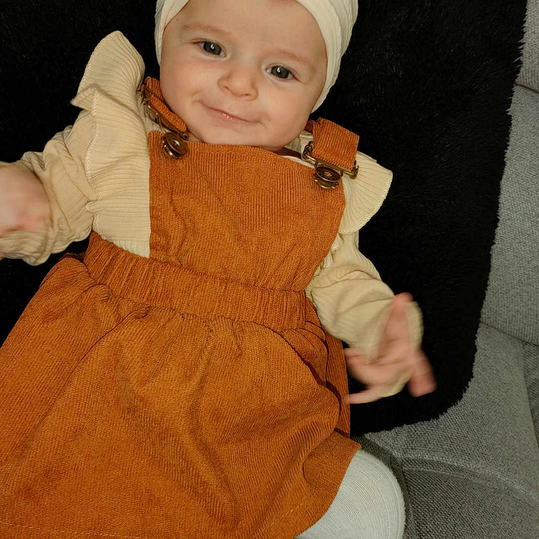 Romy a rejoint le concours — aidez-le/la à gagner de superbes lots ! baby, child, orange_dress, headband, flower, couch, black_cushion, tights, smile, cute, indoor, portrait, infant, cozy, clothing, person, baby_girl, sitting, headwear, young_child