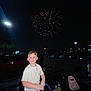 child, boy, fireworks, night, parking_lot, car, people, celebration, flash, smile, clothing, chain_necklace, dark_sky, outdoor, festive, illumination, urban, excited, standing, event