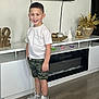 child, boy, smile, white_tshirt, camo_shorts, sneakers, indoor, floor, wooden_floor, cabinet, decor, golden_statue, vase, golden_foliage, love_sculpture, modern, furniture, happy, standing, casual