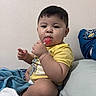 Remington is registered to the contest to win money with this photo: toddler, child, lollipop, yellow_shirt, indoor, bed, blanket, toy, pillow, wall, sitting, cute, young_child, face, hand, short_hair, casual_clothing, snack, relaxed, home