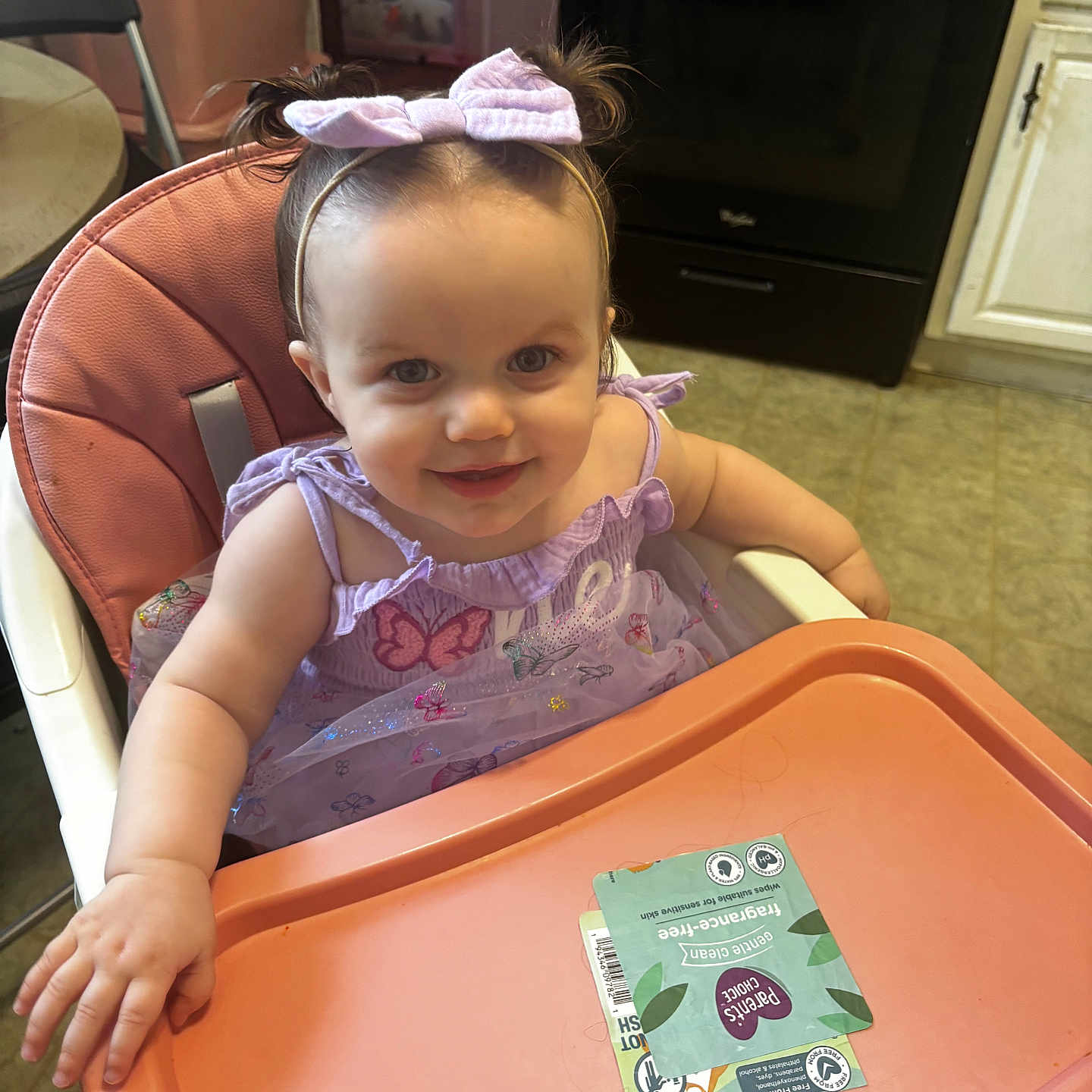 Lailah is registered to the contest to win money with this photo: baby, child, toddler, smiling, happy, portrait, high_chair, bow, headband, lavender_dress, butterfly, kitchen, indoor, tray, snack_package, home, furniture, seated, facial_expression, close_up