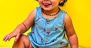 Mila Raine is registered to the contest to win money with this photo: toddler, child, baby, red_bow, blue_romper, yellow_background, sitting, cute, smiling, playful, tongue_out, jewelry, necklace, earrings, indoor, portrait, happy, barefoot, curly_hair, skin