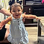 baby, black_hair, child, cute, dress, floor, furniture, hair_bows, happy, home, indoor, person, portrait, smile, sneakers, standing, supporting_hand, table, toddler, young_child
