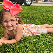 Luna is registered to the contest to win money with this photo: accessory, bow, casual_clothing, child, curly_hair, cute, daylight, face, girl, glasses, grass, greenery, happy, lying_down, nature, outdoor, park, portrait, smile, summer