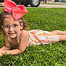 accessory, bow, casual_clothing, child, curly_hair, cute, daylight, face, girl, glasses, grass, greenery, happy, lying_down, nature, outdoor, park, portrait, smile, summer