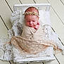 newborn, baby, swaddle, headband, flower_crown, sleeping, bed, white_bed, lace_blanket, wooden_floor, infant, cute, peaceful, portrait, resting, soft_texture, wrapped, small_bed, delicate, cozy