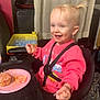 cake, child, curtain, face, food, frosting, furniture, hand, happy, high_chair, indoor, messy, person, pink_sweatshirt, plate, pony_tail, seat_belt, smiling, table, toddler