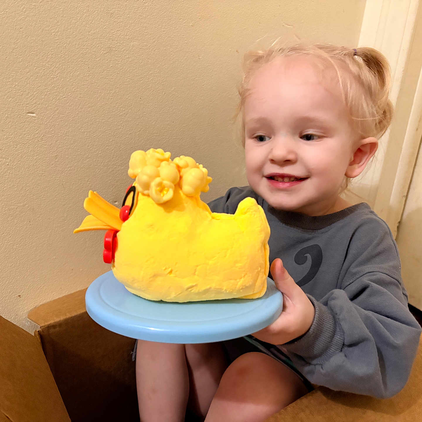 Emilia is registered to the contest to win money with this photo: toddler, child, cake, yellow_cake, duck_cake, blue_plate, cardboard_box, pigtails, smiling, happy, indoor, wall, sitting, holding, food, birthday_cake, young_child, playful, curious, light_skinned