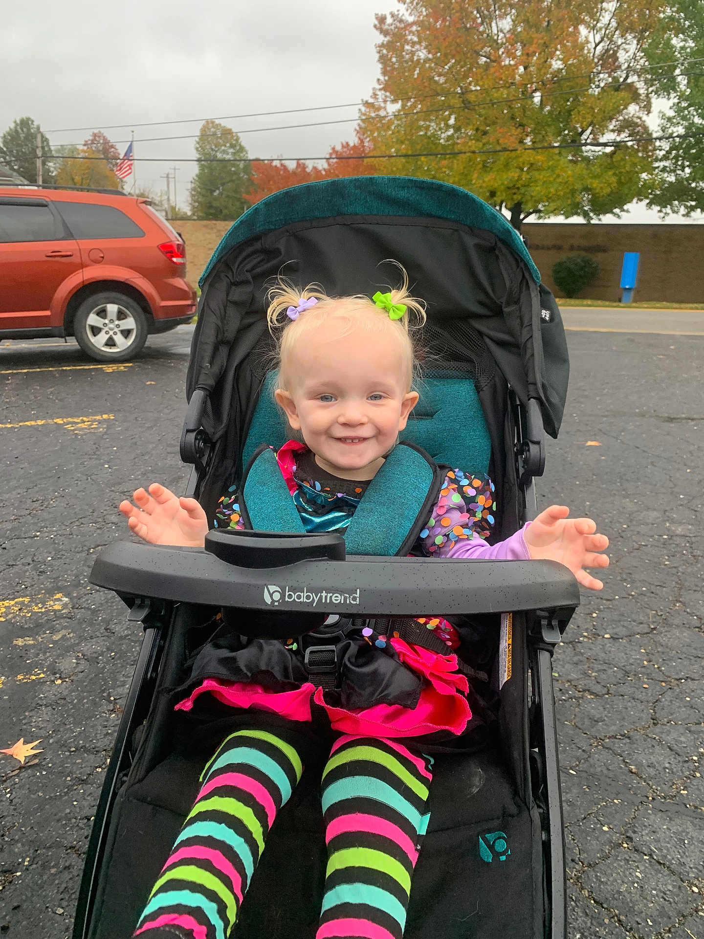 Emilia is registered to the contest to win money with this photo: toddler, stroller, child, smile, colorful_clothing, striped_leggings, autumn, fall_foliage, parking_lot, car, tree, outdoor, hair_ties, babytrend_stroller, happy, playful, pigtails, pavement, person, daytime