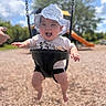 Ember is registered to the contest to win money with this photo: baby, child, swing, playground, hat, smiling, happy, outdoor, sunny, blue_sky, trees, slide, hand, clothing, shorts, tshirt, daytime, fun, recreation, park