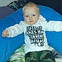 toddler, child, blue_eyes, white_shirt, text_on_shirt, camo_pants, cushion, blanket, indoor, person, baby, casual_clothing, sitting, cute, young_child, comfortable, portrait, resting, home, curious